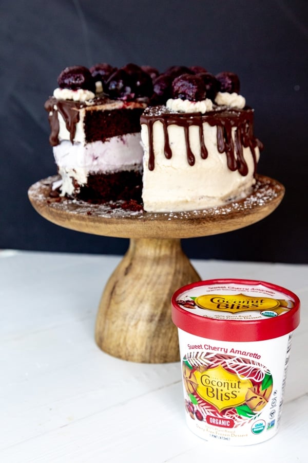 A chocolate & cherry ice cream cake with chocolate dripping down the sides and cherries on top on a wood cake stand and a pint of Coconut Bliss Cherry Amaretto ice cream next to it.