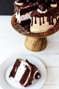 A slice of ice cream cake on a white plate with the whole cake on a wood cake stand behind it.