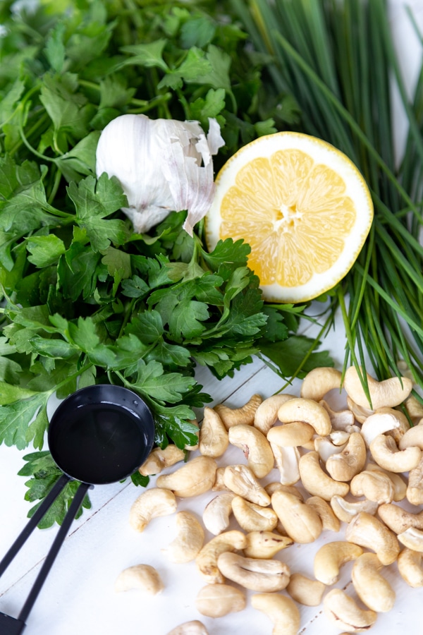 Parsley, chives, lemon, a black measuring spoon with vinegar, garlic, and cashews on a white wood table.