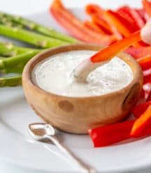 A hand dipping a slice of red pepper into a wood bowl of ranch dressing.