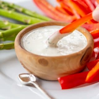 A hand dipping a slice of red pepper into a wood bowl of ranch dressing.