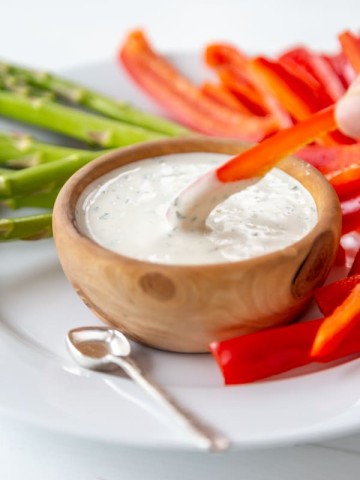 A hand dipping a slice of red pepper into a wood bowl of ranch dressing.