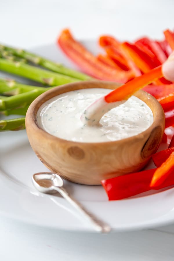 A hand dipping a slice of red pepper into a wood bowl of ranch dressing.