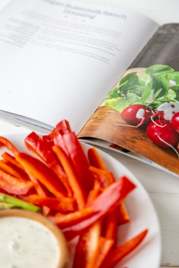 A white plate with sliced red pepper and a wood bowl with ranch dip and an open cookbook with the ranch recipe showing.