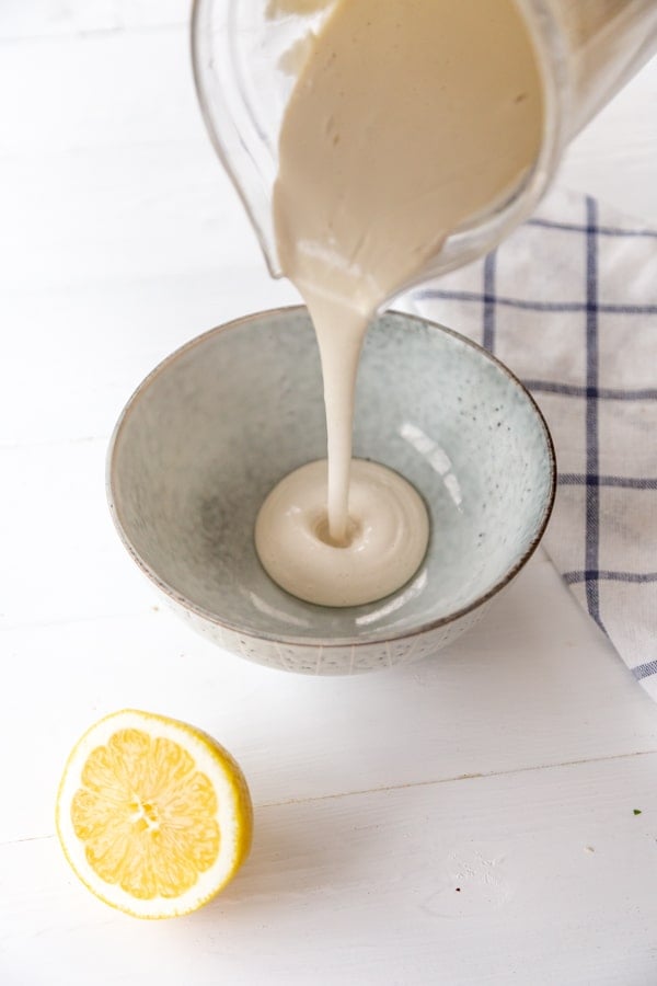 A pitcher of ranch dressing being poured into a bowl with a lemon and checked towel next to the bowl.