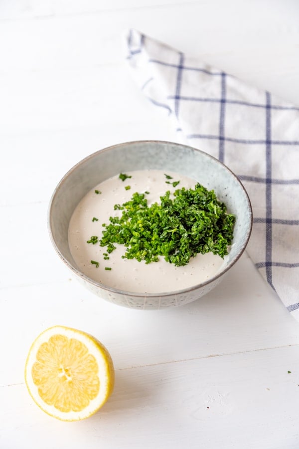 A bowl of ranch dressing with the chopped herbs sitting on top of it and a lemon and checked towel next to the bowl.