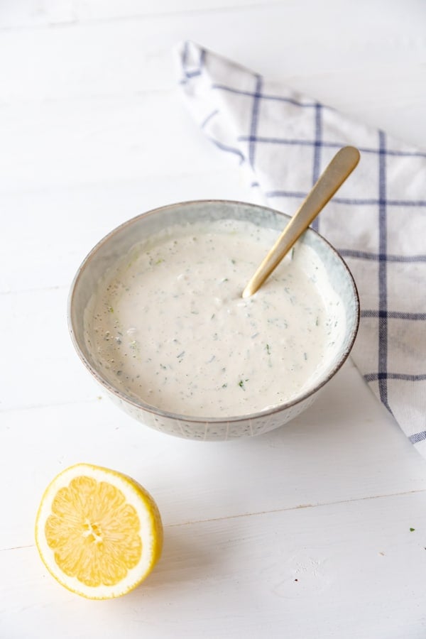 A bowl of ranch dressing with a spoon in the bowl and a half of a lemon and a checked towel next to the bowl.