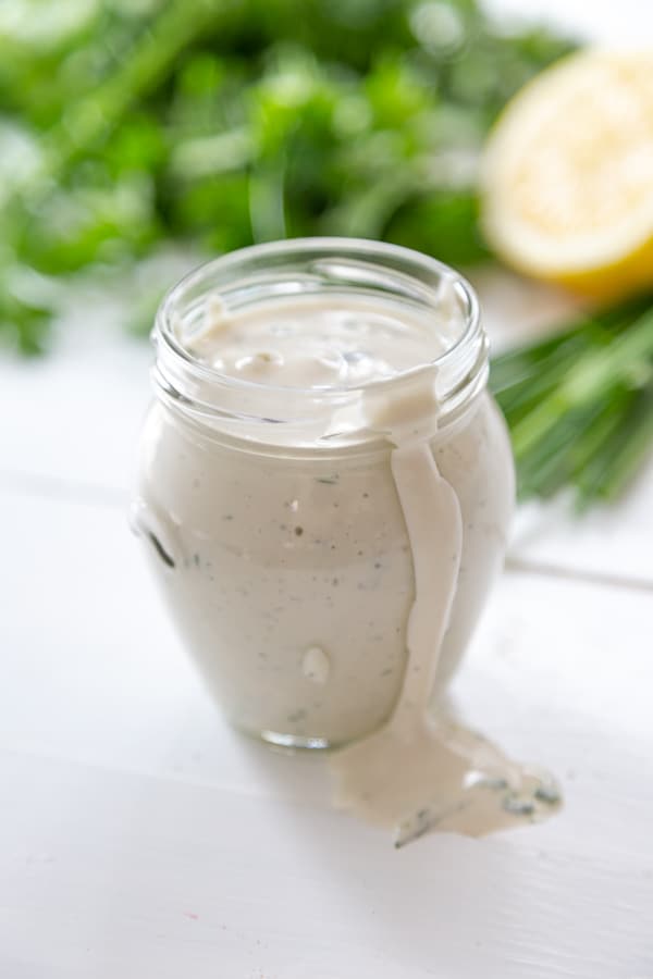 A glass jar filled with ranch dressing with some dripping down the sides and herbs and lemon in the background.