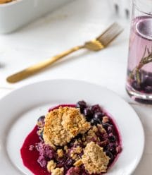 A blueberry crisp on a white plate with the pan of crisp in the background.