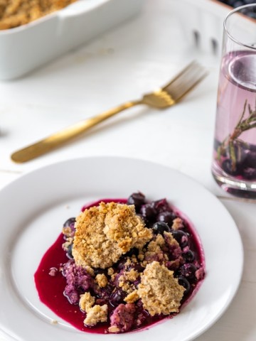A blueberry crisp on a white plate with the pan of crisp in the background.