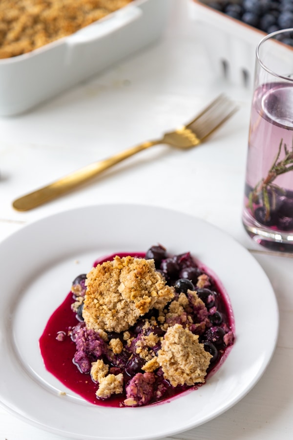 A blueberry crisp on a white plate with the pan of crisp in the background.