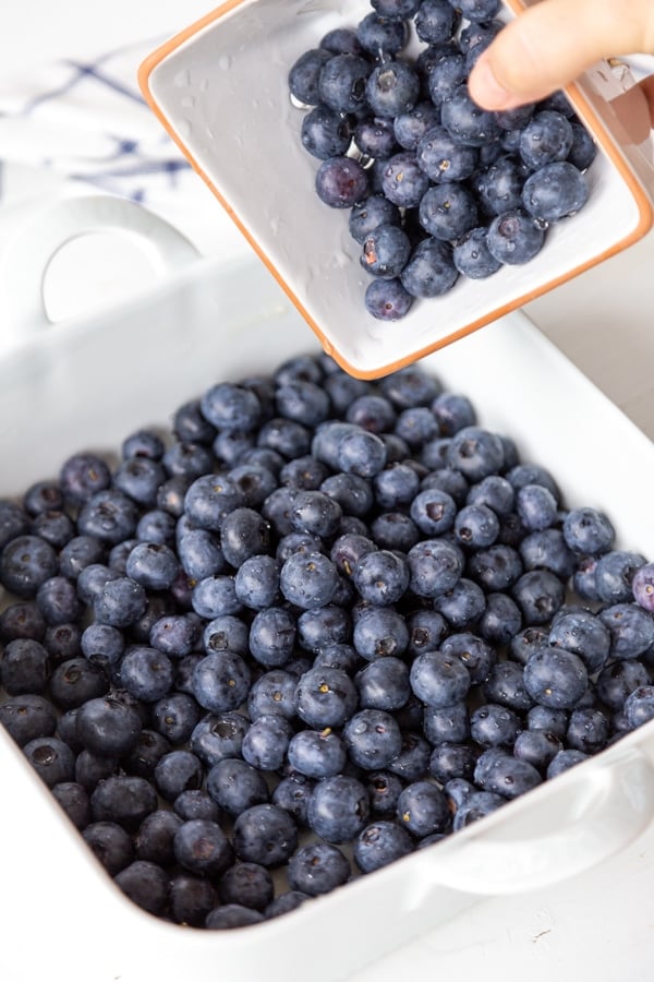 A hand holding pouring a crate of blueberries into a white pan.