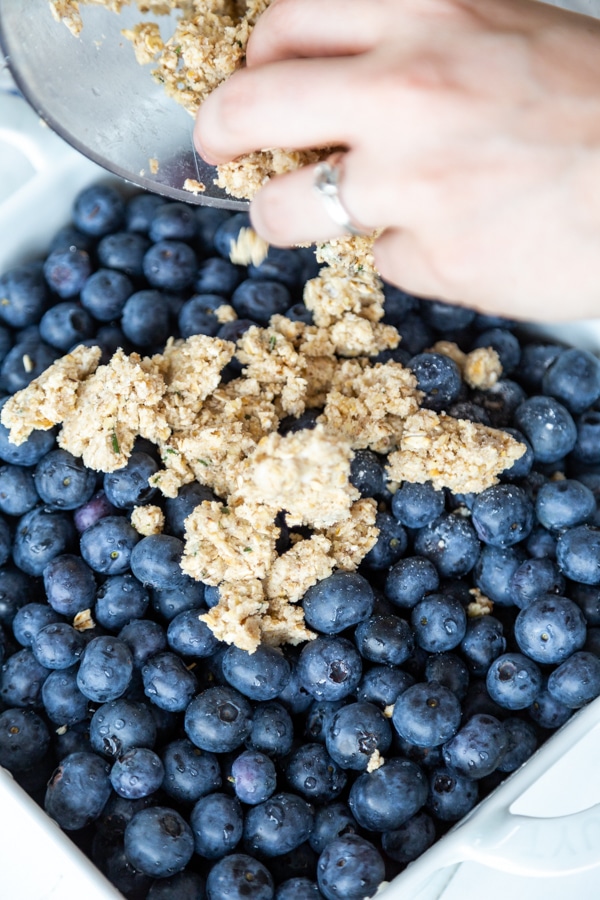 A hand sprinkling an oat and flour topping over a pan of fresh blueberries.