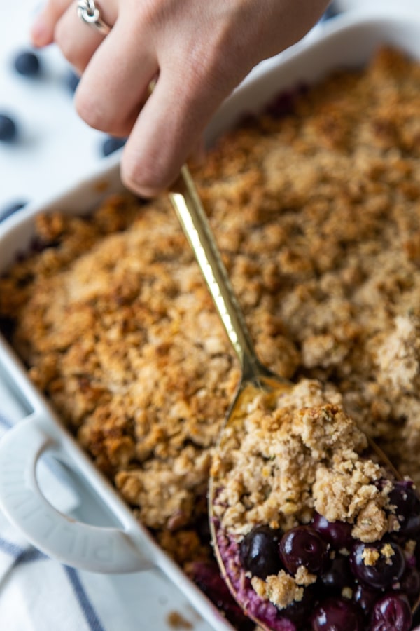 A hand holding a spoon, scooping out a serving of blueberry crumble from a pan.