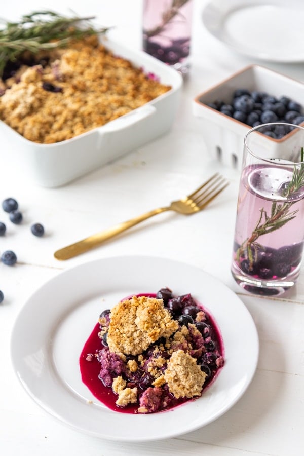 A tablescape with a plate of blueberry crisp and a white pan of the crisp with a glass of blueberry water.