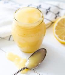 A glass jar of lemon curd with a silver spoon dripping curd next to it and a half of a lemon.