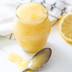 A glass jar of lemon curd with a silver spoon dripping curd next to it and a half of a lemon.