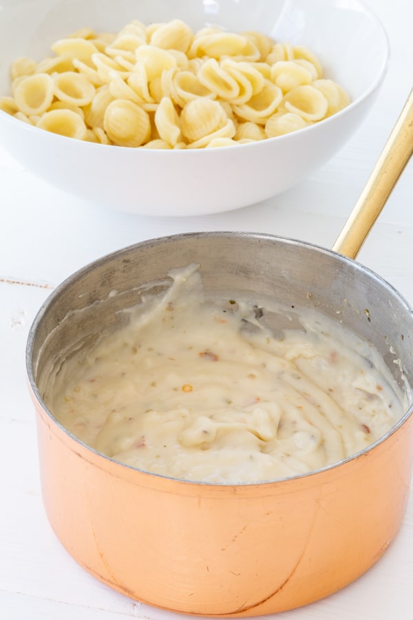 A copper pan with white cream sauce and a bowl of pasta next to it.