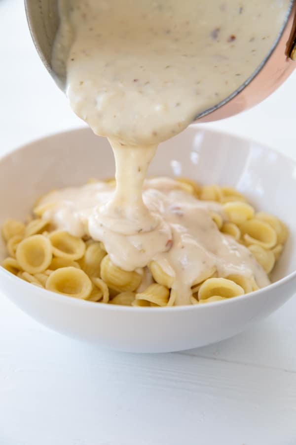 Pasta in a white bowl with a pan of cream sauce being poured on top of it.