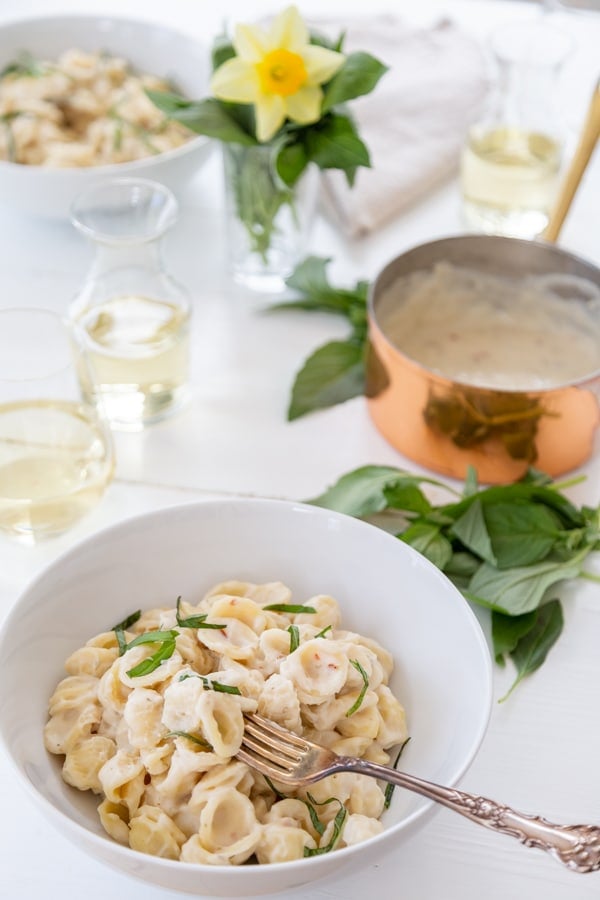 A white bowl of pasta with cream sauce and a copper pot of sauce and basil and lemons on the table. 