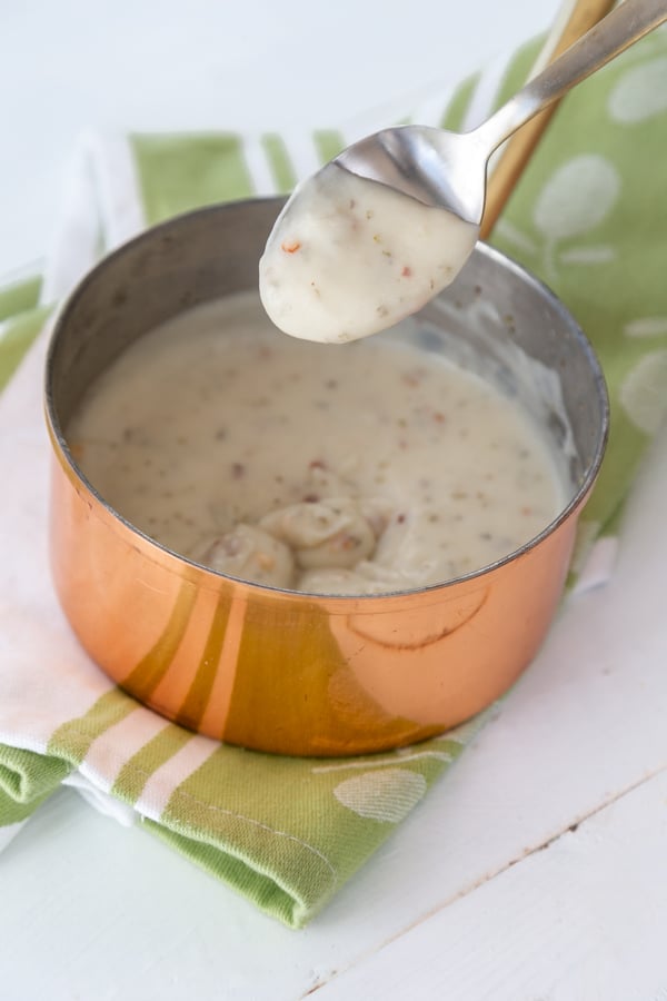 A spoon with cream sauce hanging over a copper pan of white cream sauce.