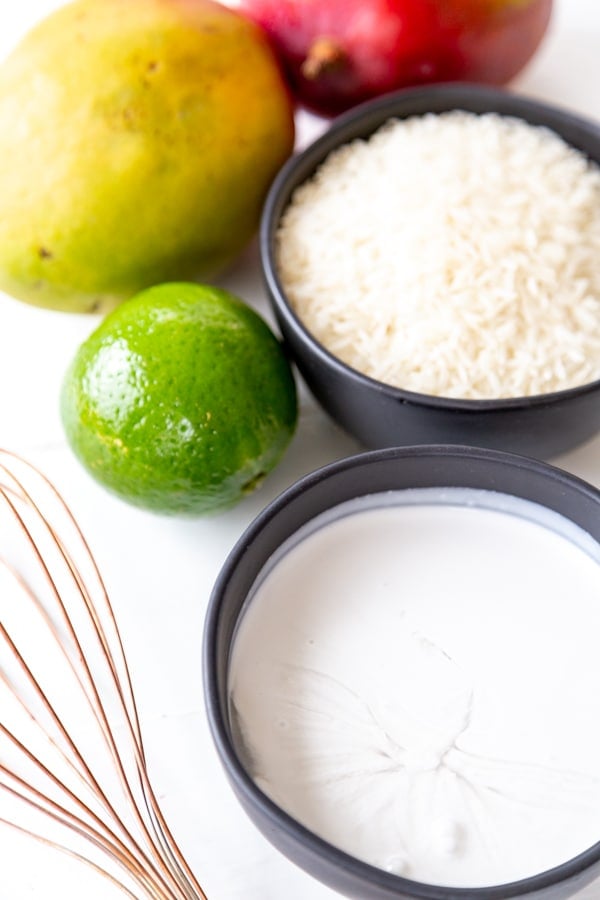 A bowl of milk, rice, a lime, mangos, and a copper whisk on a white table.