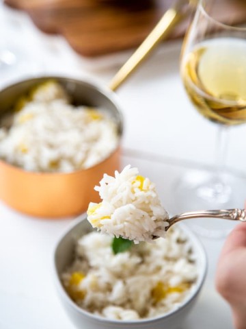 A hand holding a fork with mango rice over a bowl of rice and a copper pot of rice and a glass of wine in the background.