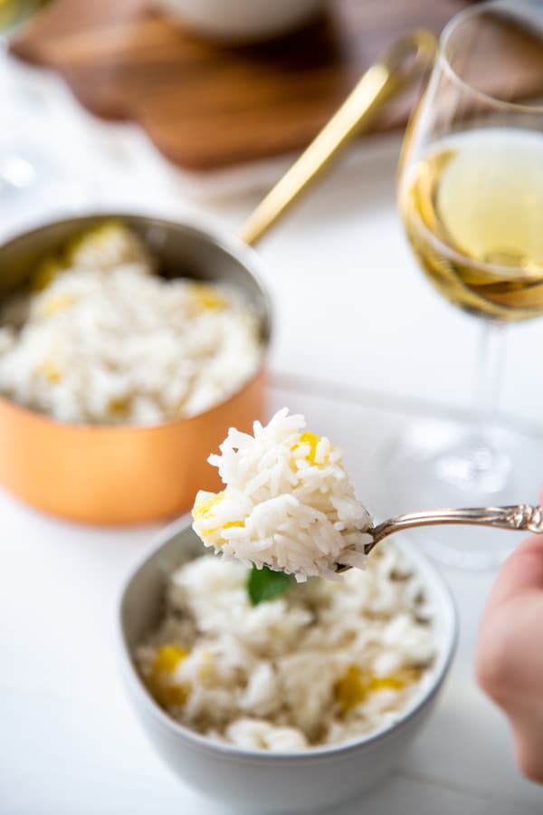 A hand holding a fork with mango rice over a bowl of rice and a copper pot of rice and a glass of wine in the background. 