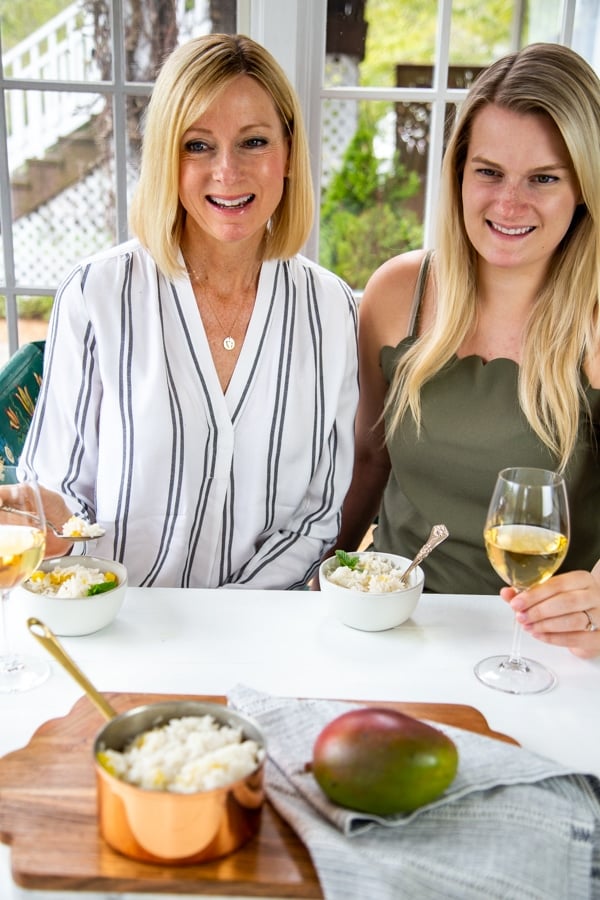 Two blonde women smiling and eating a bowl of rice and drinking white wine.