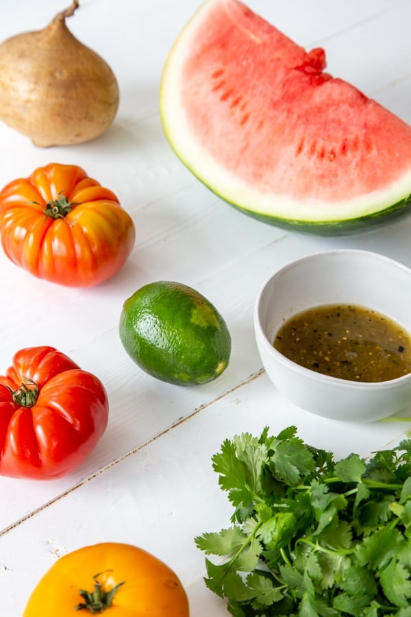 A slice of watermelon, heirloom tomatoes, a whole jicama, cilantro, and a white bowl of tomatilla salsa