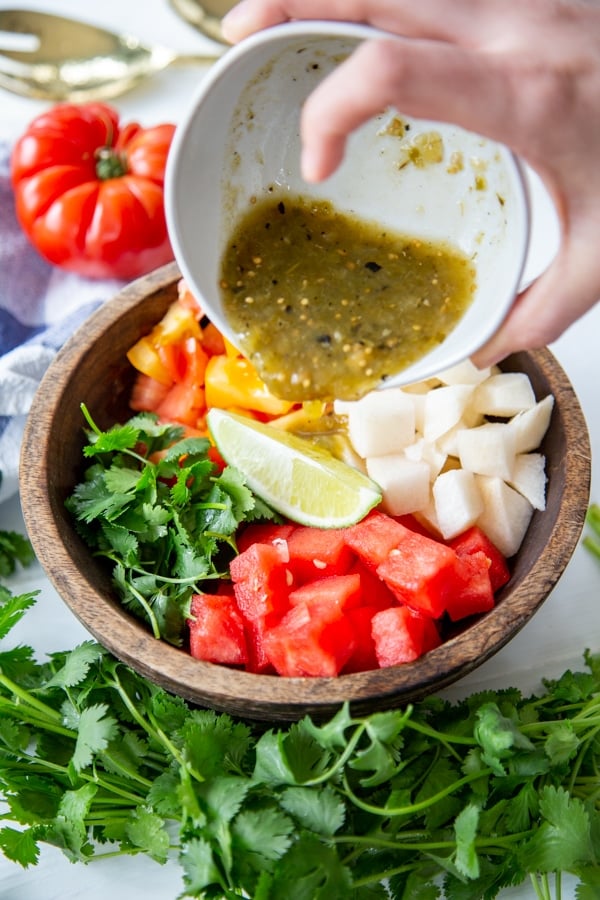 A hand pouring a bowl of salsa on a watermelon salad.