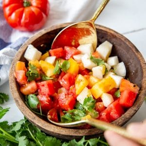 A wood bowl with hands holding gold spoons and tossing a watermelon salad.