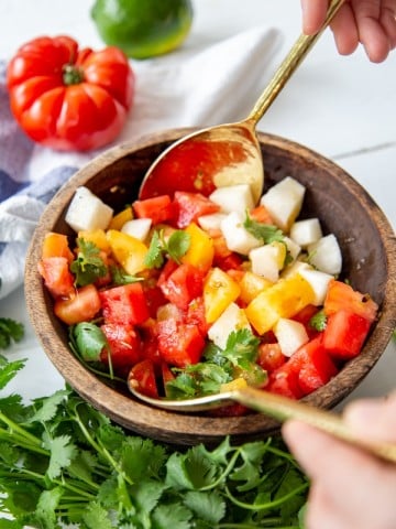 A wood bowl with hands holding gold spoons and tossing a watermelon salad.