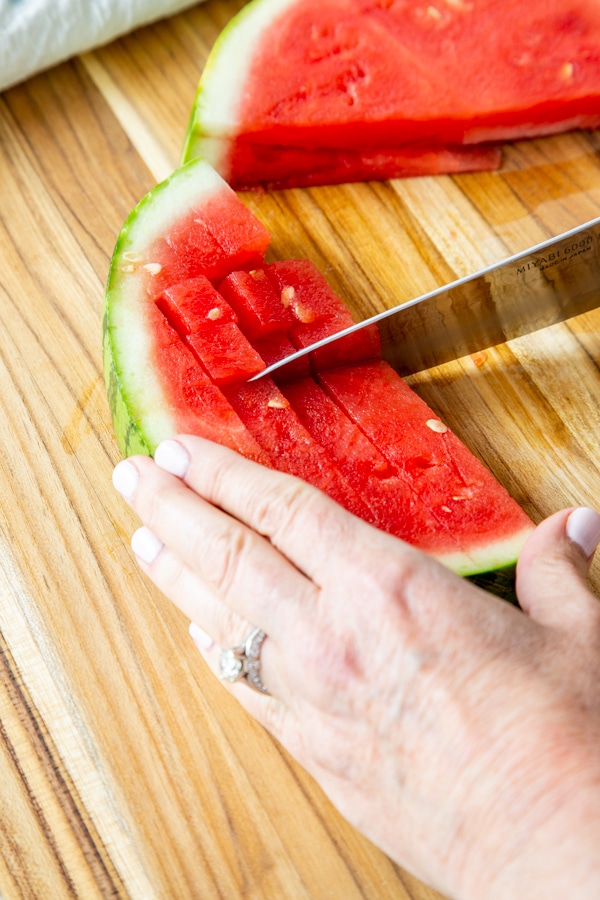 A hand with a sharp knife cubing a watermelon.