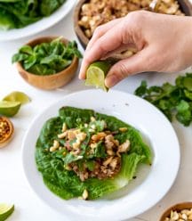 A hand squeezing a lime wedge over a vegetable lettuce wrap on a white plate with bowls of ingredients next to it.