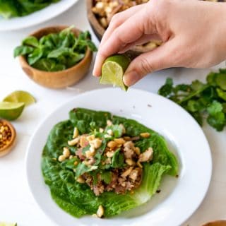 A hand squeezing a lime wedge over a vegetable lettuce wrap on a white plate with bowls of ingredients next to it.