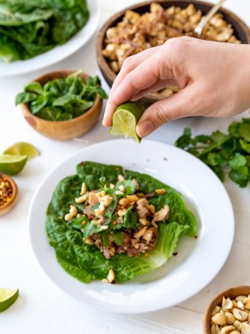 A hand squeezing a lime wedge over a vegetable lettuce wrap on a white plate with bowls of ingredients next to it.
