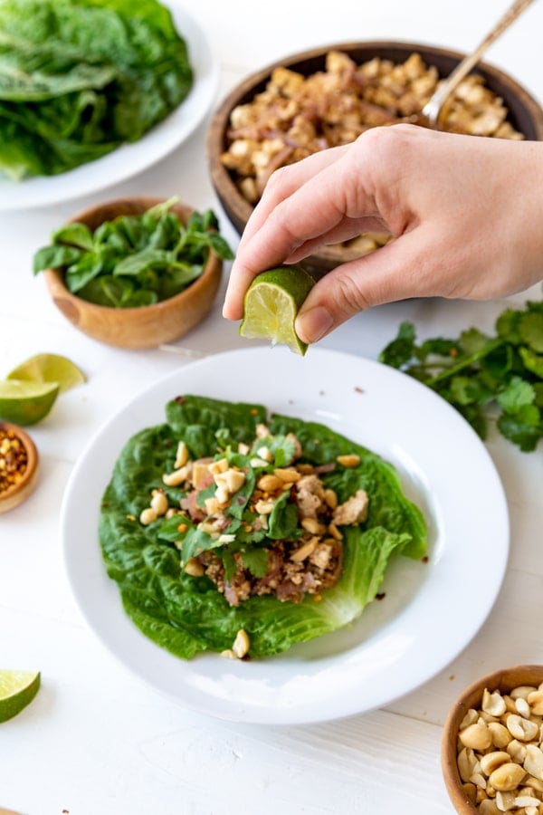 A hand squeezing a lime wedge over a vegetable lettuce wrap on a white plate with bowls of ingredients next to it. 