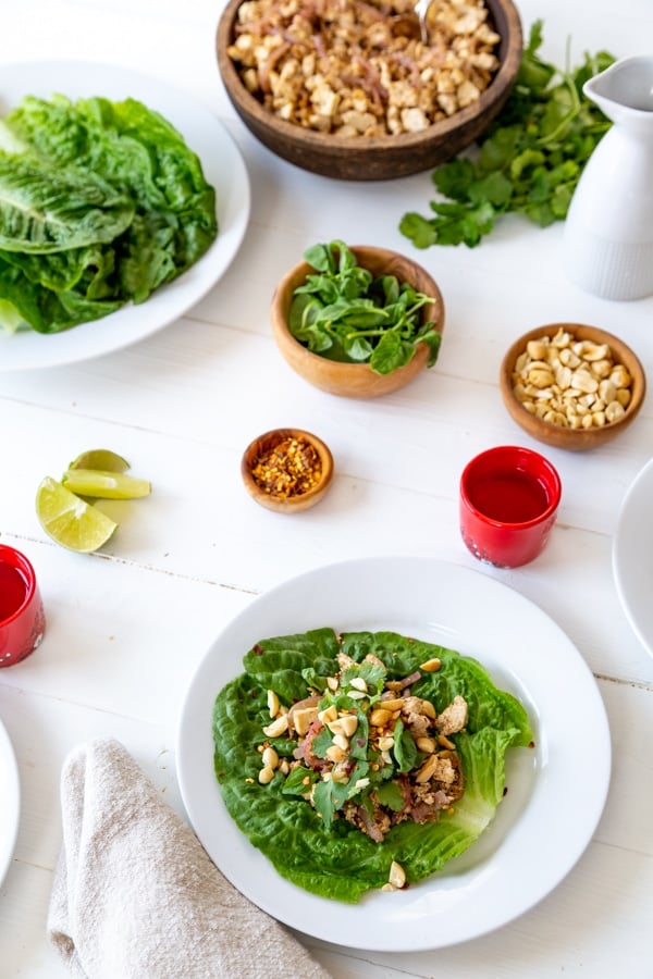 A tablescape of lettuce wraps on white plates and bowls of ingredients on a white wood table. 