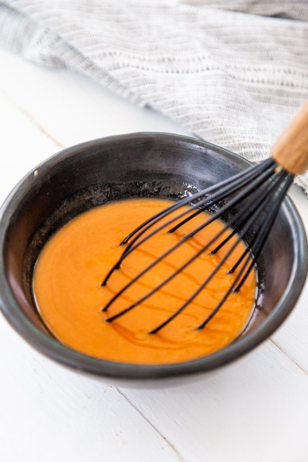 Buffalo sauce in a black bowl with a black and wood whisk in the bowl. 