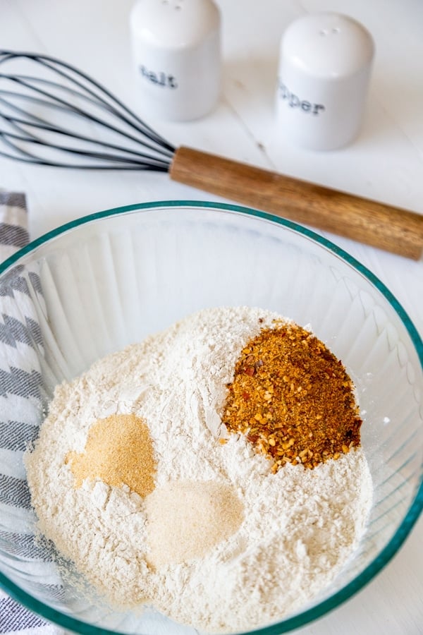 A glass bowl with flour and spices and a wooden whisk next to it.