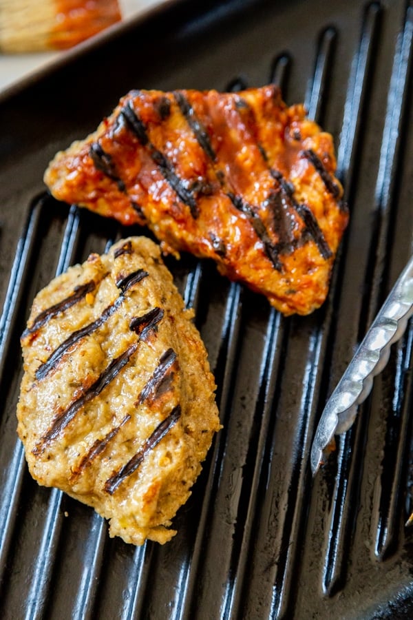 Two pieces of grilled chik'n on a grill pan,, one with BBQ sauce.