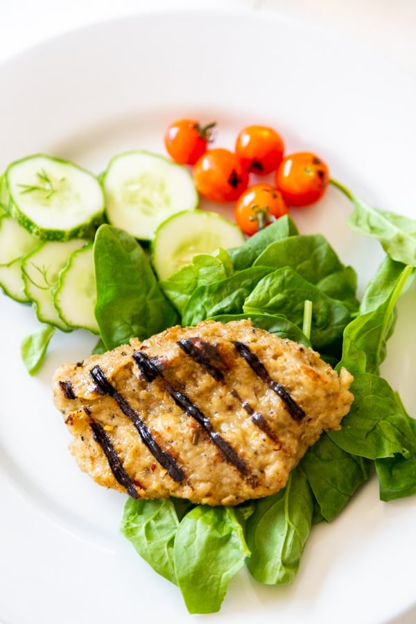 A grilled chicken breast on a bed of spinach with tomatoes and cucumbers on a white plate. 