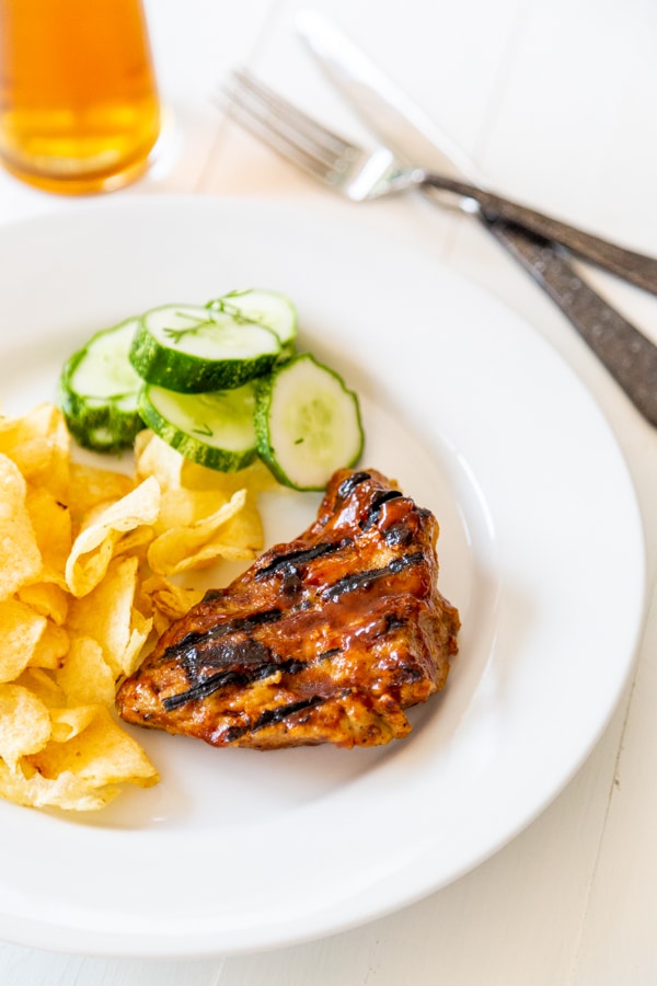 Grilled chicken, chips, and cucumbers on a white plate with a glass of beer and utensils. 