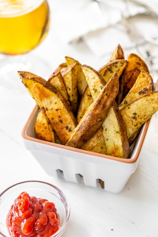 Home fries in a white dish with a small dish of ketchup and a glass of beer next to it. 