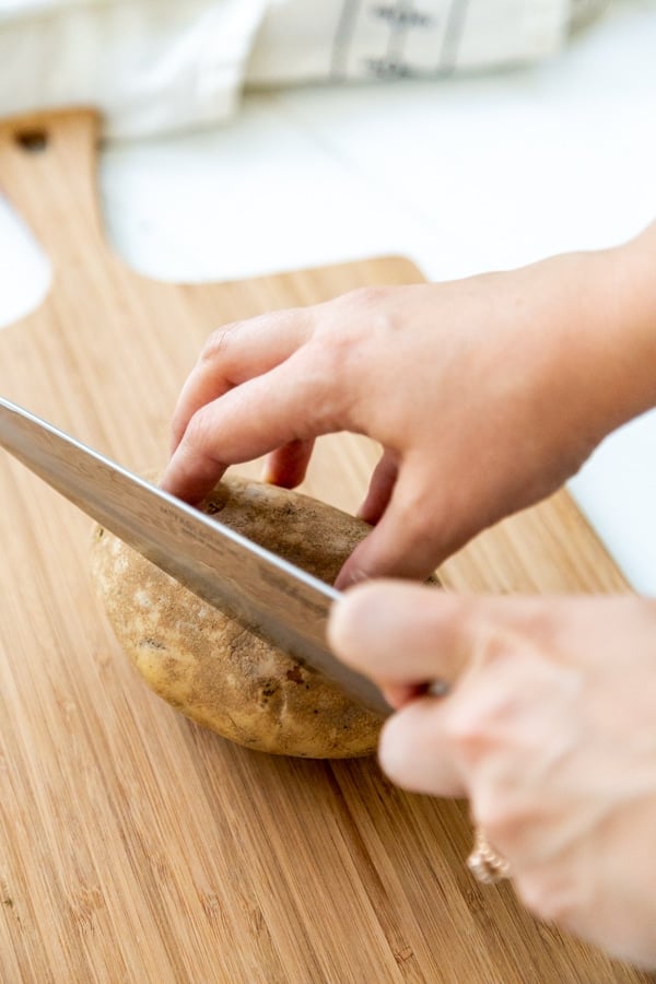 A hand slicing a potato with a knife on a wood cutting board.