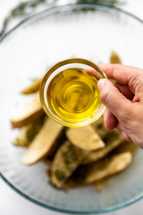 A hand holding a dish of olive oil over sliced potatoes.