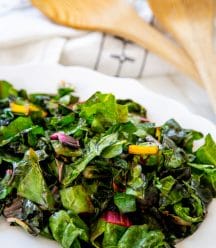 A white plate with sauteed rainbow chard and a wood serving spoon and fork in the background.