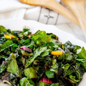 A white plate with sauteed rainbow chard and a wood serving spoon and fork in the background.