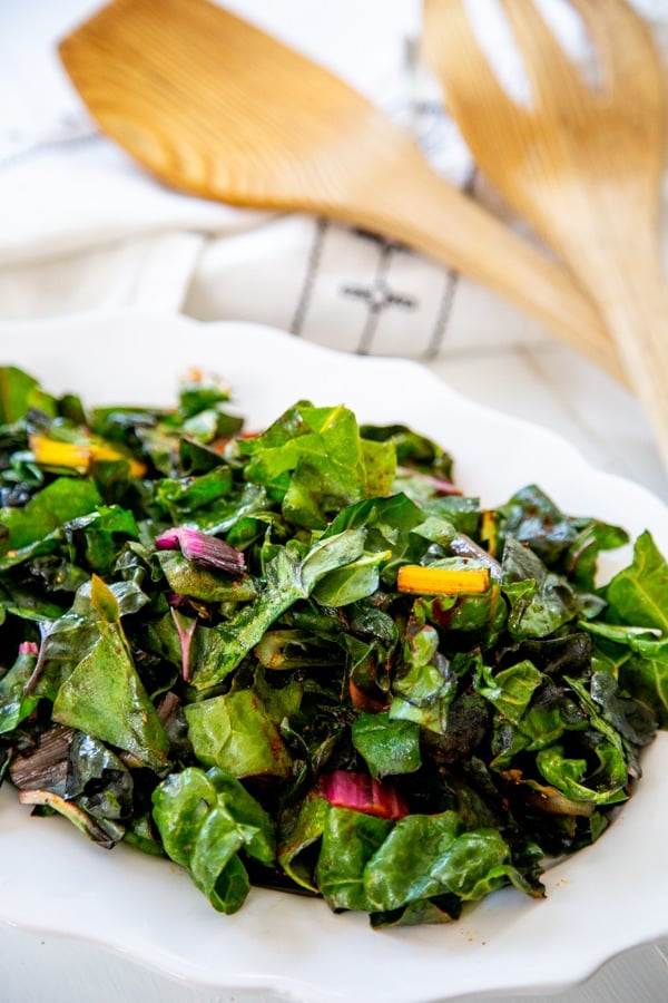 A white plate with sauteed rainbow chard and a wood serving spoon and fork in the background.